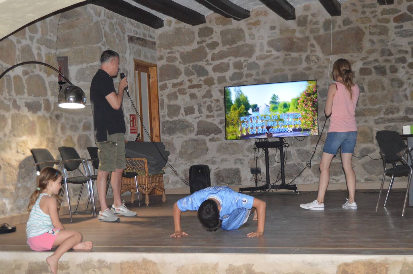 Imatge on es veuen uns adults cantant en un karaoke i uns nens jugant dins una sala del Castell de Pradell, allotjament privat per a grups.